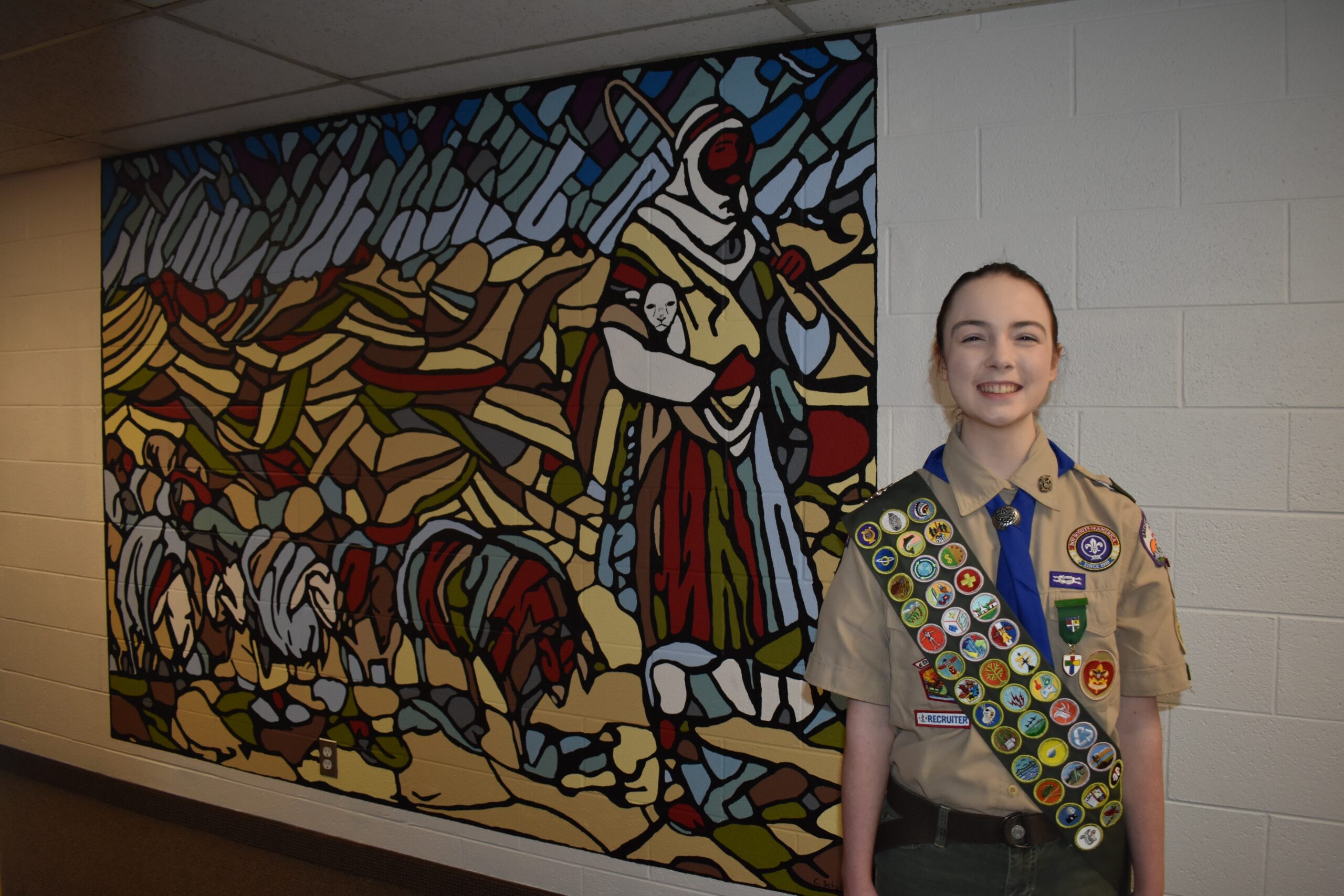 Eagle Scout Chloe Schultz Kellett standing next to a mural of a shepard leading sheep.